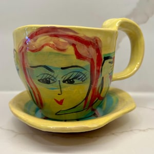 Könnte beinhalten: Ein handbemaltes Keramik-Tee-Tassen- und Untertassen-Set. Die Tasse und die Untertasse sind gelb, mit einem bemalten Gesichtsdesign mit roten Haaren, blauem Lidschatten und einer roten Lippe. Der Griff ist gebogen.