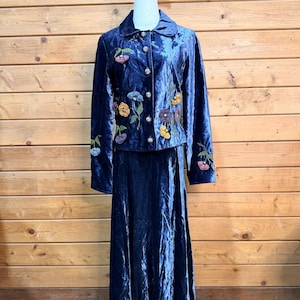 May include: A dark blue velvet two-piece set consisting of a long-sleeved button-down jacket and a matching floor-length skirt. The jacket features embroidered floral motifs in yellow, purple, and orange, with silver-toned metallic buttons.