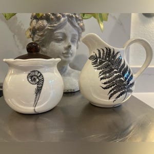 Puede incluir: Un azucarero de cerámica blanca con tapa marrón oscuro y una cremaera a juego. Ambas piezas presentan un diseño de helecho negro. El azucarero tiene un diseño de helecho en espiral, mientras que la cremaera tiene una hoja de helecho completa.
