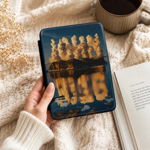 May include: A blue and black tablet case featuring a landscape image of a lake reflecting clouds and mountains. The case is held in a person's hand, with a white sweater sleeve visible. A cup of coffee and a book are in the background.