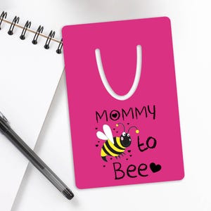Könnte beinhalten: Rosafarbener Lesezeichen mit einer Bienenillustration und dem Text "Mommy to Bee" mit einem Herz.