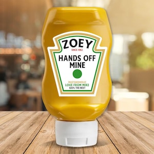 May include: A yellow mustard bottle with a white cap. The label reads "ZOEY SINCE 1982 HANDS OFF MINE" with a green dot and the text "MUSTARDHOLIC LOVE FROM MIKE 100% THE BEST". The bottle is on a wooden surface.