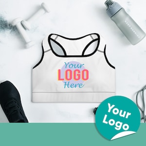 May include: White sports bra with black straps and a colorful "Your Logo Here" design. The bra is on a white surface with a water bottle and a jump rope.