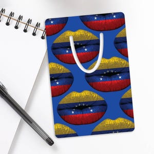 May include: A blue bookmark with a repeating pattern of lips in the colours of the Venezuelan flag: red, yellow, and blue. The bookmark has a white handle and is on a white surface next to a spiral notebook and a pen.