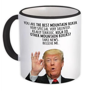 Puede incluir: Taza de cerámica blanca con asa y borde negros. La taza presenta un mensaje humorístico sobre el ciclismo de montaña y un retrato de un hombre haciendo un gesto con la mano de 'ok'. El texto dice: 'ERES EL MEJOR CICLISTA DE MONTAÑA...CRÉEME.'