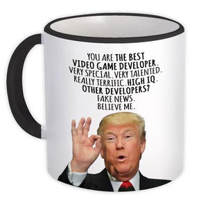 May include: White ceramic mug with a black handle and rim. The mug features a portrait and text: "YOU ARE THE BEST VIDEO GAME DEVELOPER. VERY SPECIAL. VERY TALENTED. REALLY TERRIFIC. HIGH IQ. OTHER DEVELOPERS? FAKE NEWS. BELIEVE ME."