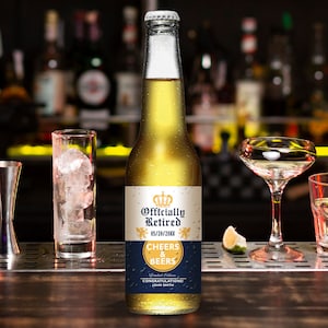 May include: A beer bottle with a custom label that reads "Officially Retired" with a crown graphic. The label also includes the date "05/20/20XX" and the words "Cheers & Beers". The bottle is on a bar with a glass of ice and a cocktail glass.
