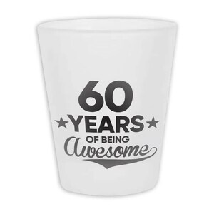 May include: A white shot glass with the text "60 YEARS OF BEING Awesome" in black. The number 60 is large and bold, with the word "YEARS" between two stars. The phrase "OF BEING Awesome" is in a script font.