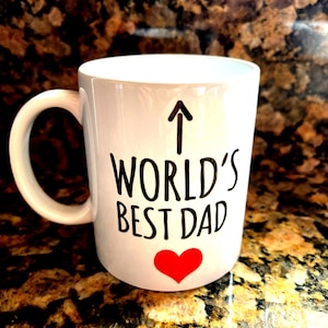 May include: White ceramic mug with a black arrow pointing up, the words "WORLD'S BEST DAD" in black, and a red heart. The mug has a curved handle and is likely used for beverages.