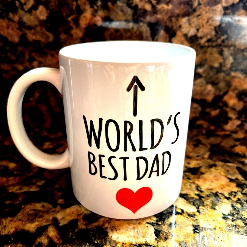 May include: White ceramic mug with a black arrow pointing up, the words "WORLD'S BEST DAD" in black, and a red heart. The mug has a curved handle and is likely used for beverages.