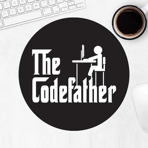 Op de afbeelding: Zwarte ronde muismat met de witte tekst "The Codefather" en een afbeelding van een persoon achter een computer. Een kopje koffie en een toetsenbord zijn ook zichtbaar.