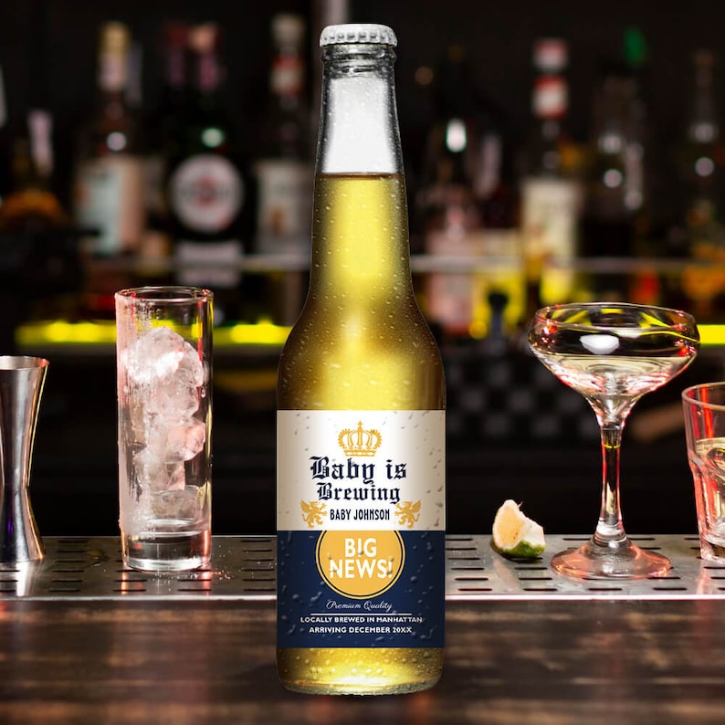May include: A clear glass bottle with a gold liquid, featuring a label that reads "Baby is Brewing" with "Big News!" and "Baby Johnson." The bottle is on a bar with glasses and a lime wedge.