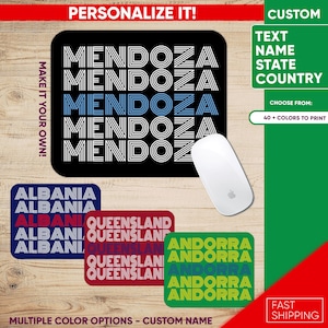 Puede incluir: Un mousepad rectangular negro con el nombre "MENDOZA" en letras blancas y azules. También se muestran mousepads adicionales con los nombres "ALBANIA", "QUEENSLAND" y "ANDORRA". Un ratón de ordenador blanco está al lado del mousepad.