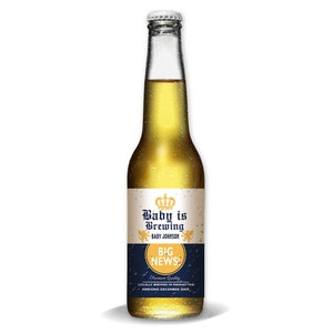 May include: A clear glass beer bottle filled with a golden liquid. The label reads "Baby is Brewing" with a crown graphic and "BIG NEWS!". The bottle has a silver cap and is against a white background.