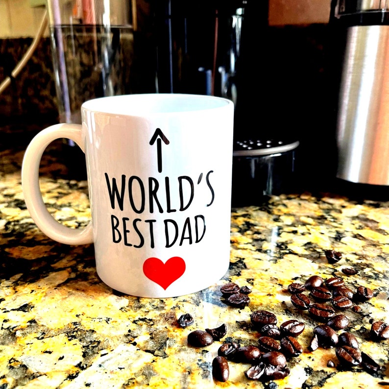 May include: White ceramic mug with the text "WORLD'S BEST DAD" in black, an upward arrow, and a red heart. Coffee beans are scattered on a speckled countertop near the mug. The mug has a curved handle.