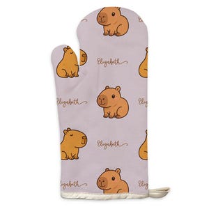 Peut inclure: Un gant de cuisine lavande avec un motif répété de capybaras de dessin animé dans des tons bruns. Le nom "Elizabeth" est écrit en cursive entre les illustrations de capybaras. Le gant a une bordure blanche et une boucle pour l'accrocher.