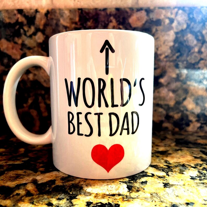 May include: White ceramic mug with the text "WORLD'S BEST DAD" in black, an upward-pointing arrow, and a red heart. The mug has a curved handle and is likely used for beverages.