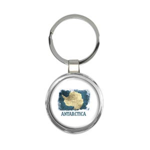 May include: A round silver keychain with a map of Antarctica in gold and blue. The word "ANTARCTICA" is printed in blue below the map. The keychain is a travel-themed accessory.