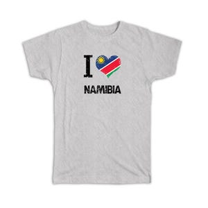 May include: A light grey short-sleeved t-shirt featuring a black letter I, a heart-shaped graphic filled with the blue, red, green, white, and yellow flag of Namibia, and the word NAMIBIA in black distressed capital letters below the heart.