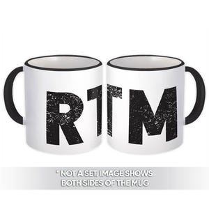 Puede incluir: Una taza de cerámica blanca con un borde y un asa negros. La taza tiene la palabra "RTM" impresa en negro con un aspecto desgastado y vintage. La imagen muestra ambos lados de la taza.
