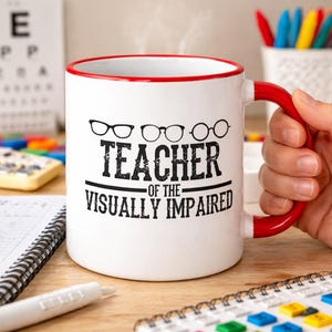 Könnte beinhalten: Weiße Keramik-Tasse mit rotem Innenraum und Henkel. Die Tasse trägt den schwarzen Text "TEACHER OF THE VISUALLY IMPAIRED" sowie drei Brillenpaare. Die Tasse steht auf einer Holzoberfläche.