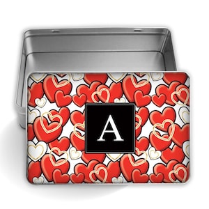 May include: A silver metal tin with a white background and a red heart pattern. The letter "A" is in a black square in the center of the tin.