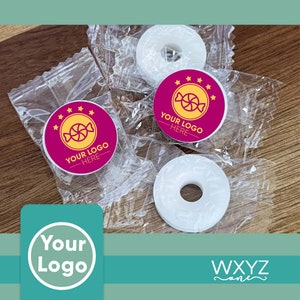 Op de afbeelding: Drie witte ronde muntjes met een gat in het midden, verpakt in doorzichtige plastic zakjes. Elk zakje heeft een roze en gele sticker met de tekst "YOUR LOGO HERE" en een snoepillustratie.