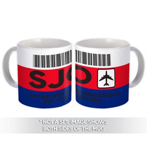 May include: White ceramic mug with a gray handle, featuring a red, white, and blue design. The mug displays the letters "SJO" in black, along with a barcode and an airplane icon. Text on the mug reads "Juan Santamaria San Jose" and "International Airport Costa Rica."
