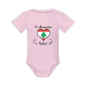 May include: A pale pink baby bodysuit with short sleeves. The front displays a heart-shaped design with the Lebanese flag and the words "Lebanese Baby!" in black script. The garment is made of a soft material.