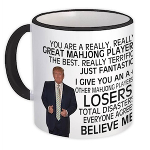 Op de afbeelding: Een witte keramische mok met een zwarte rand. De mok heeft een zwart-witte afbeelding van een man in een zwart pak en een rode stropdas. De tekst op de mok luidt: "You are a really, really great Mahjong player the best. Really terrific just fantastic I give you an A+ Other Mahjong players Losers Total disasters Everyone agree Believe me"