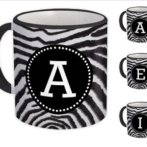 May include: A ceramic coffee mug with a black handle and rim, featuring a zebra stripe pattern. The mug displays the letter "A" in white within a black circle. Additional mugs with the letters "E" and "I" are also shown.
