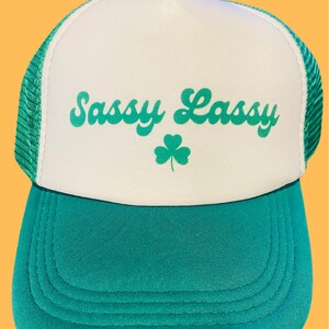 May include: White and green trucker hat with the text "Sassy Lass" in green cursive lettering and a green shamrock.