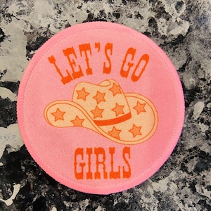 Puede incluir: Parche redondo rosa con el texto "Let's Go Girls" en naranja. Un sombrero de vaquero de dibujos animados con estrellas naranjas está en el centro del parche.