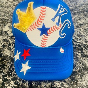 May include: A blue trucker hat with a white baseball, a gold crown, and the letters "KC" embroidered on the front. The hat also has two stars on the side, one red and one white.