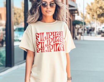 May include: Cream-colored t-shirt with the word "Filipina" in red, stacked three times. The shirt has short sleeves and a relaxed fit. The person is wearing sunglasses and a watch.