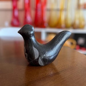 May include: A black ceramic bird figurine with etched leaf and swirl designs. The bird is perched on a wooden surface. The figurine has a smooth, glossy finish and is a decorative item.
