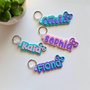 May include: Four personalized name keychains in various colors. Each keychain features a name in a playful font, with a bow accent. The names are Olivia, Kaia, Sophia, and Fiona. The keychains are on a white surface.