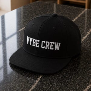 Op de afbeelding: Een zwarte honkbalpet met de tekst VYBE CREW geborduurd in witte hoofdletters op het voorpaneel. De pet heeft een gestructureerde kroon, een platte klep en kleine ventilatiegaatjes op de bovenste panelen van de hoed.