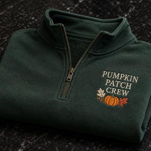 Puede incluir: Una sudadera verde oscuro con cremallera de un cuarto y un diseño bordado en el pecho. El bordado incluye el texto PUMPKIN PATCH CREW en letras blancas sobre un gráfico de una calabaza naranja rodeada de hojas de otoño de colores.