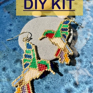 Pode incluir: Kit DIY para fazer brincos de beija-flor. Os brincos são feitos com contas coloridas e fio dourado. Os brincos são mostrados em uma superfície de pedra cinza.