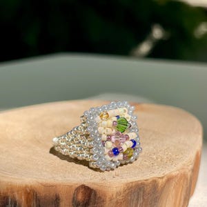 May include: A handmade beaded ring with a square top. The ring has a silver band and a square top decorated with cream, pink, green, and blue beads. The ring is displayed on a wooden surface.