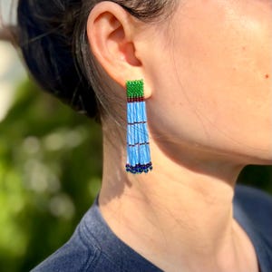 May include: A long, dangling earring crafted from small glass beads. The top section is a rectangular block of green beads, followed by five vertical strands of light blue beads, accented with small dark red and dark blue bead details.