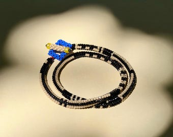 May include: A beaded bracelet featuring a repeating pattern of black and beige seed beads. The bracelet includes a toggle clasp made of bright blue square beads and a single cylindrical beige bead with yellow accents at each end.