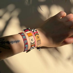 May include: A collection of beaded bracelets in various colors and patterns. One bracelet features a teal and burgundy design, while others showcase orange, black, and white geometric patterns. The bracelets are worn together, adding a vibrant touch to the wrist.