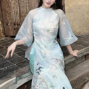 May include: A light blue, long dress with a floral and bird design. The dress has sheer, flared sleeves and a high neckline. The design includes white flowers and blue birds on branches. A white headband completes the look.