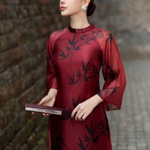 May include: A woman wearing a red silk dress with a black bamboo print. The dress has a high neck and long sleeves. She is holding a black and silver fan.