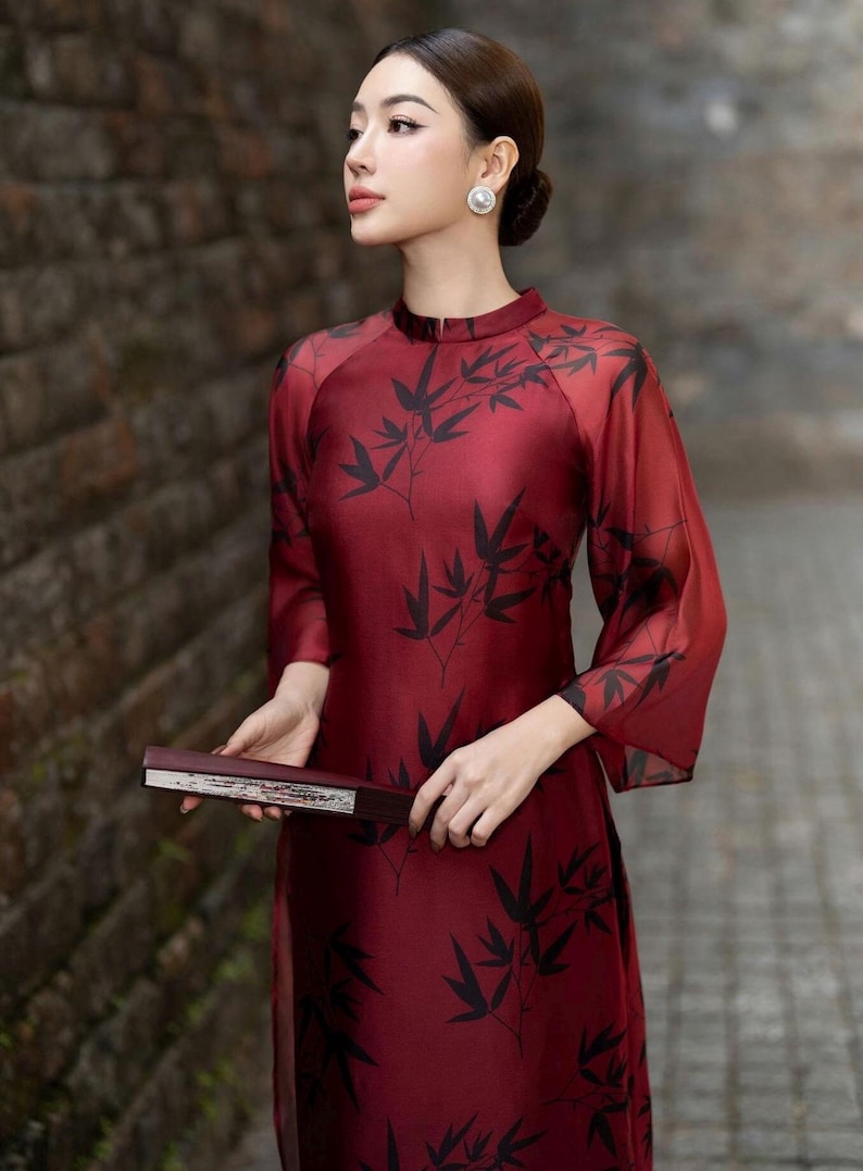 May include: A woman wearing a red silk dress with a black bamboo print. The dress has a high neck and long sleeves. She is holding a black and silver fan.