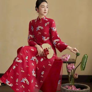 May include: A red, long-sleeved dress with a mandarin collar and floral embroidery. The dress features a high slit and is paired with a fan. The image also includes a floral arrangement in a stone bowl.
