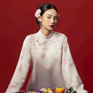 May include: A woman wearing a light pink, long-sleeved dress with a floral and butterfly pattern. The dress has a mandarin collar and frog closures. The background is a solid red. The woman has flowers in her hair and is wearing earrings.