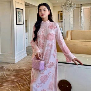 May include: A woman wearing a pink and white floral dress with a mandarin collar. She is holding a small lavender quilted handbag with a chain strap.